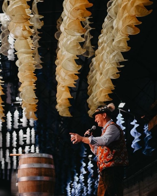 Jason Hurta in Bavarian costume on stage with a microphone beside a wooden keg, under hanging festival decorations