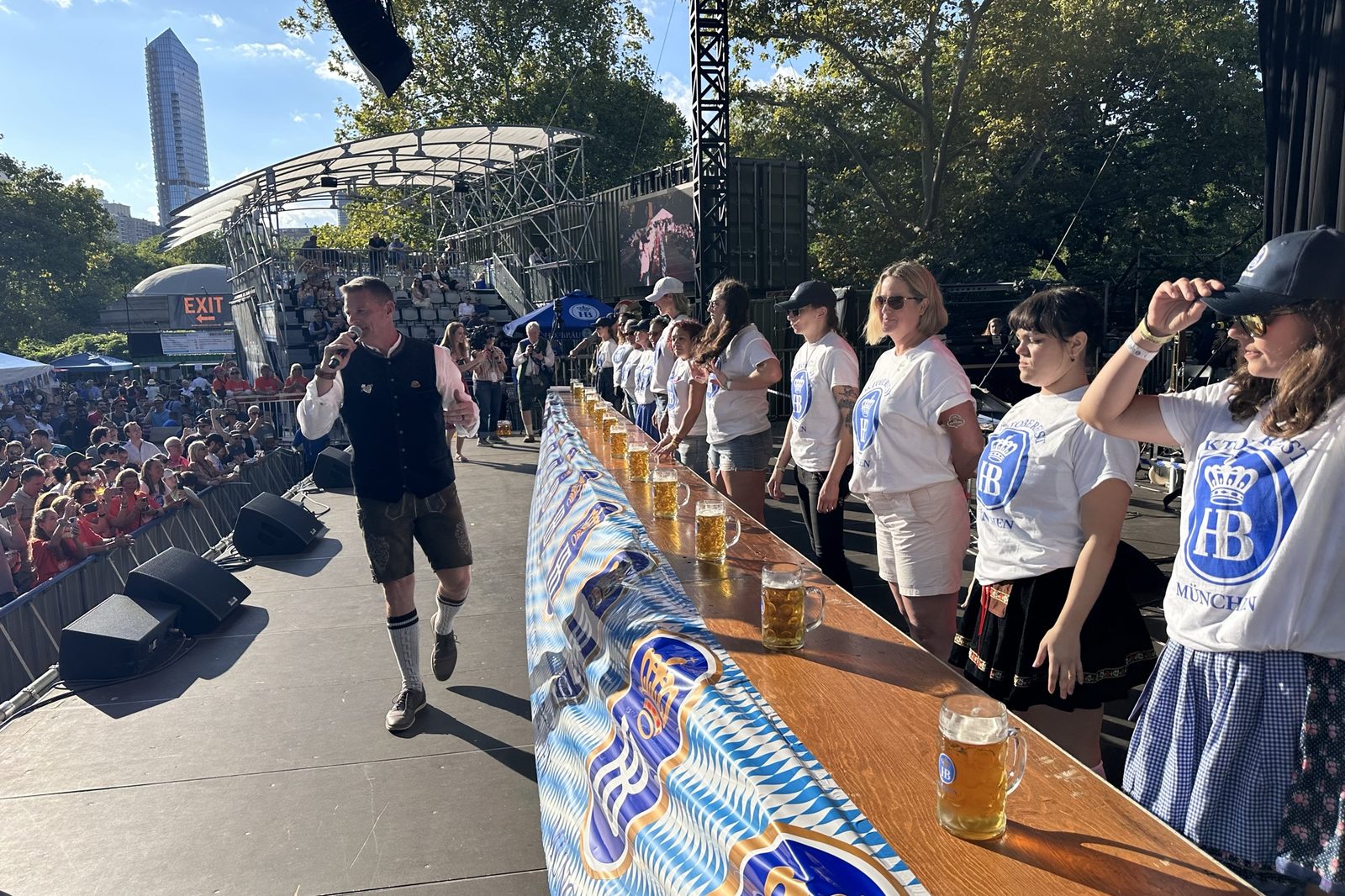 Jason Hurta calling a stein-holding competition on stage in Central Park as competitors line up behind full steins